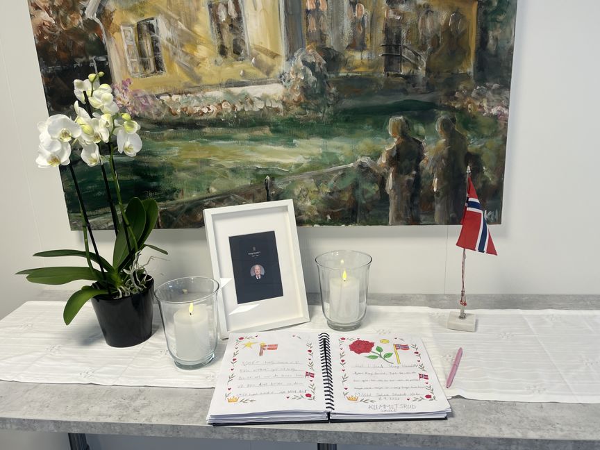 Foto: Kondolanseprotokollen fra Klemetsrud skole og AKS Et minnebord med innrammet bilde av H.M. kong Harald V, blomster, tente lys og en kondolanseprotokoll på